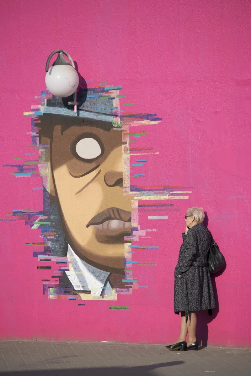 Older woman standing beside a giant mural face on a bright pink wall